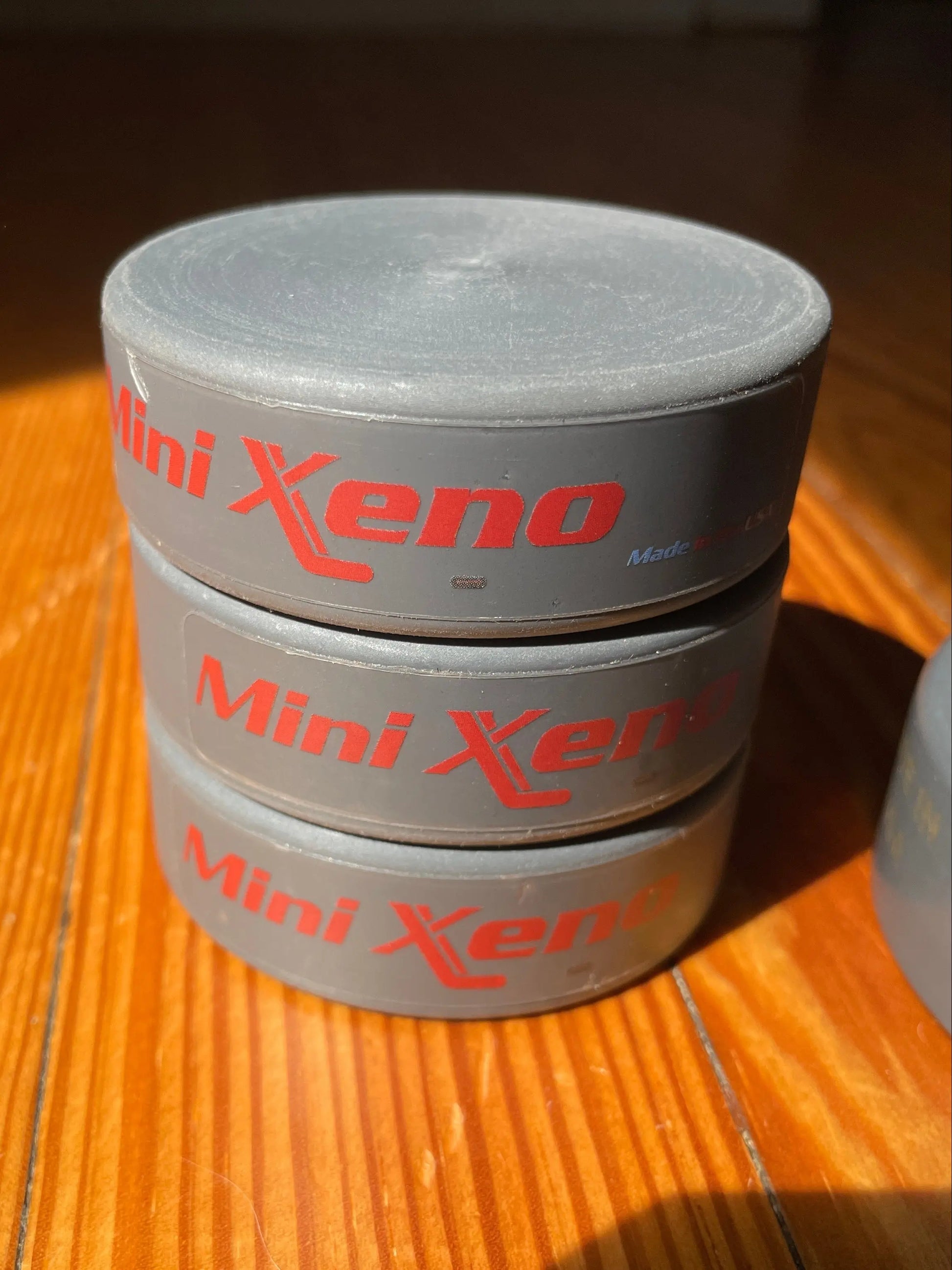Three gray Mini Xeno hockey pucks stacked vertically on a wooden table under direct sunlight.