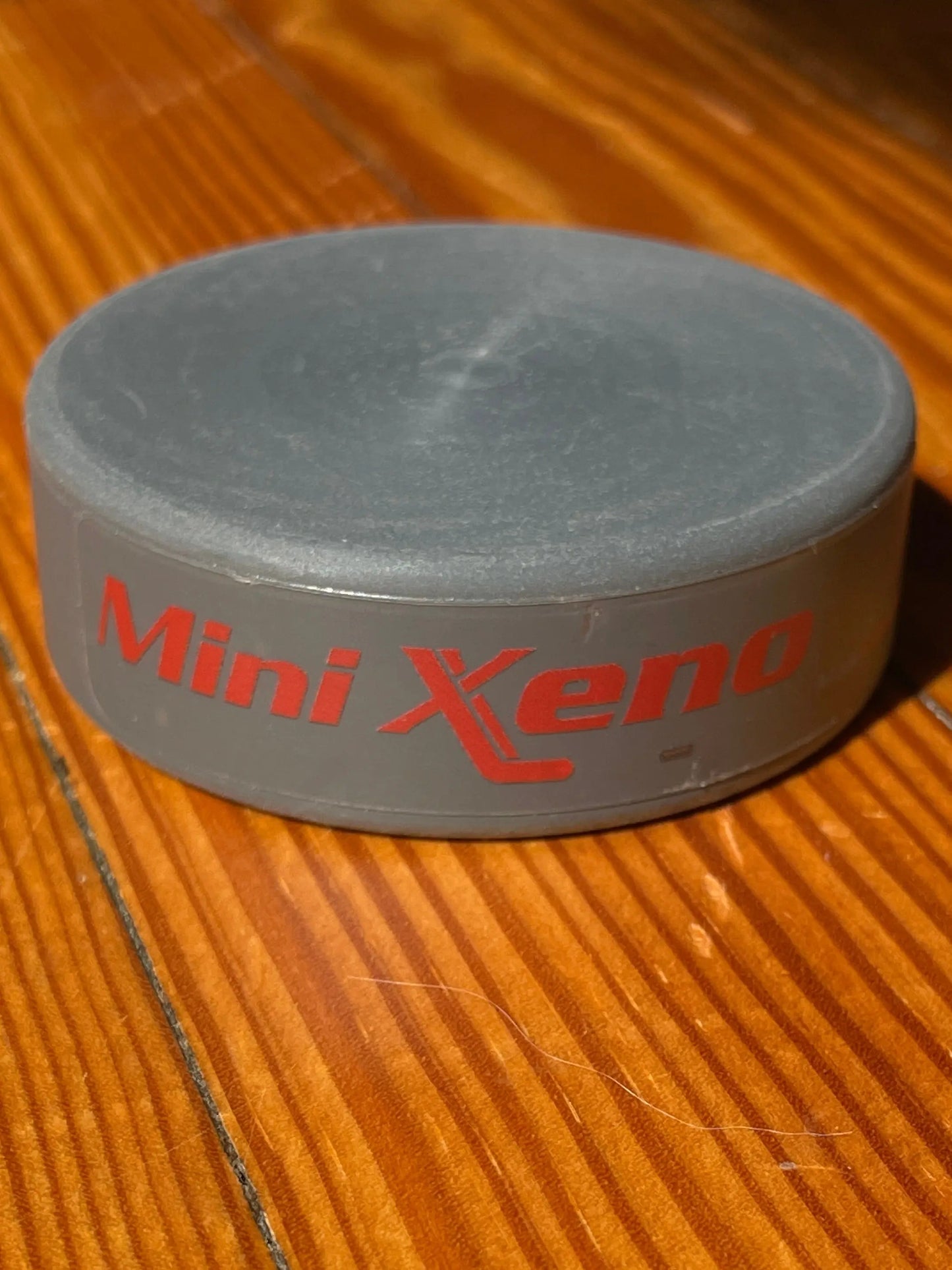 A gray Mini Xeno puck item with red 'Mini Xeno' text, photographed on a warm wood surface.