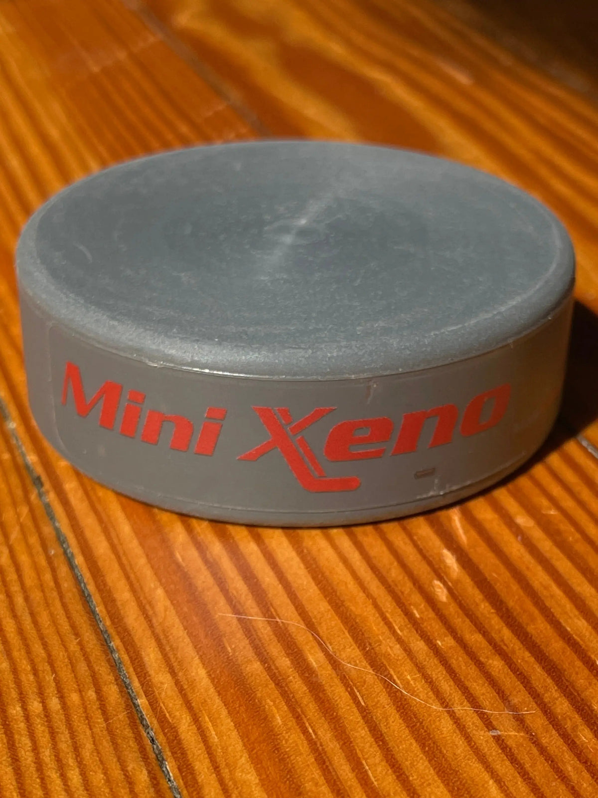 A gray Mini Xeno puck item with red 'Mini Xeno' text, photographed on a warm wood surface.