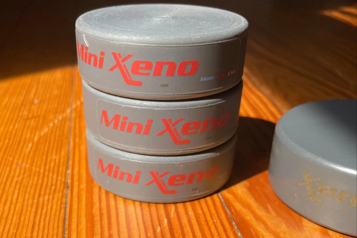 Three round gray Mini Xeno  stacked on a wooden table, with a traditional Xenopuck partially visible on the right. The pucks are photographed in direct sunlight on a wooden surface.
