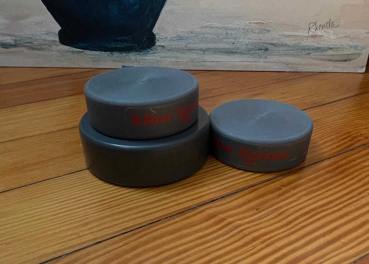 Three dark gray hockey pucks with red cursive writing, stacked and standing on a wooden floor in front of a painting. 
Xenopuck comparison shows Mini Xenos' are smaller than regular Xenopucks. 