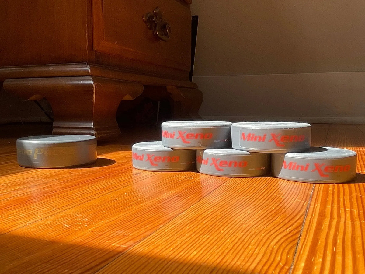 Stacked Mini Xenos' next to a regular Xenopuck on a polished wooden floor in warm sunlight, with a wooden cabinet in the background.